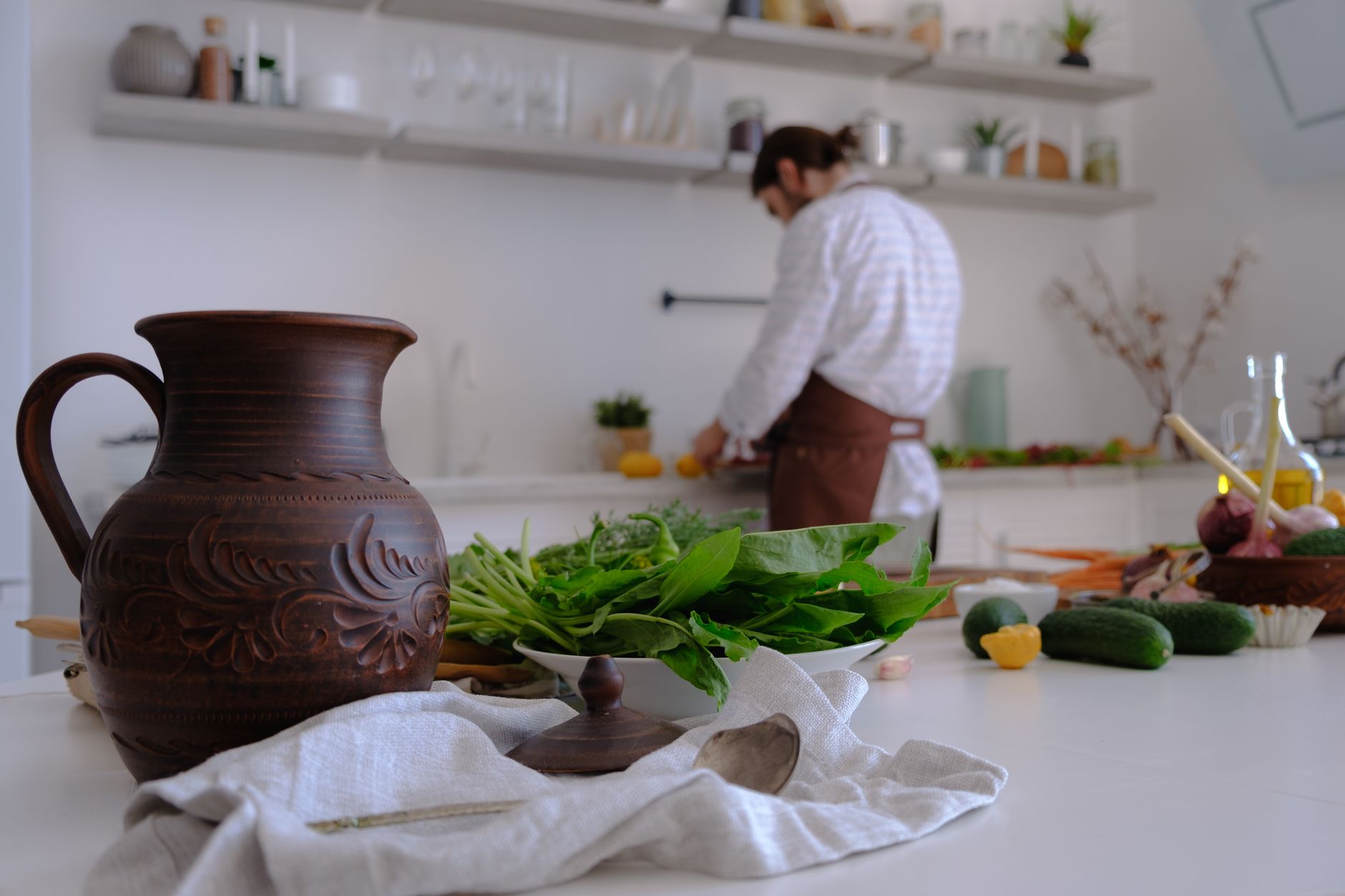 A man prepares fresh ingredients in a bright, rustic kitchen with a focus on a decorative jug and greens.